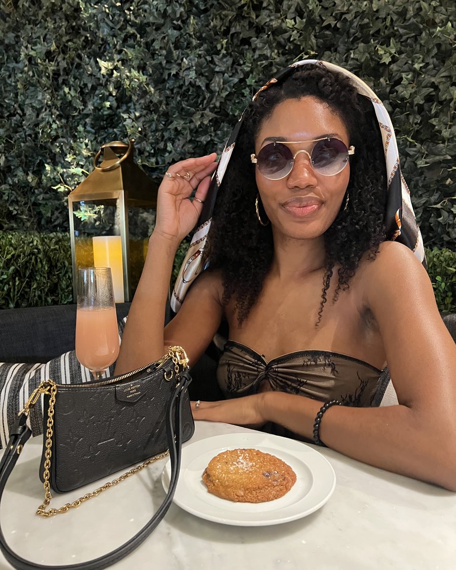 Ruqayyah seated at an outdoor table, sunglasses on, bag beside her