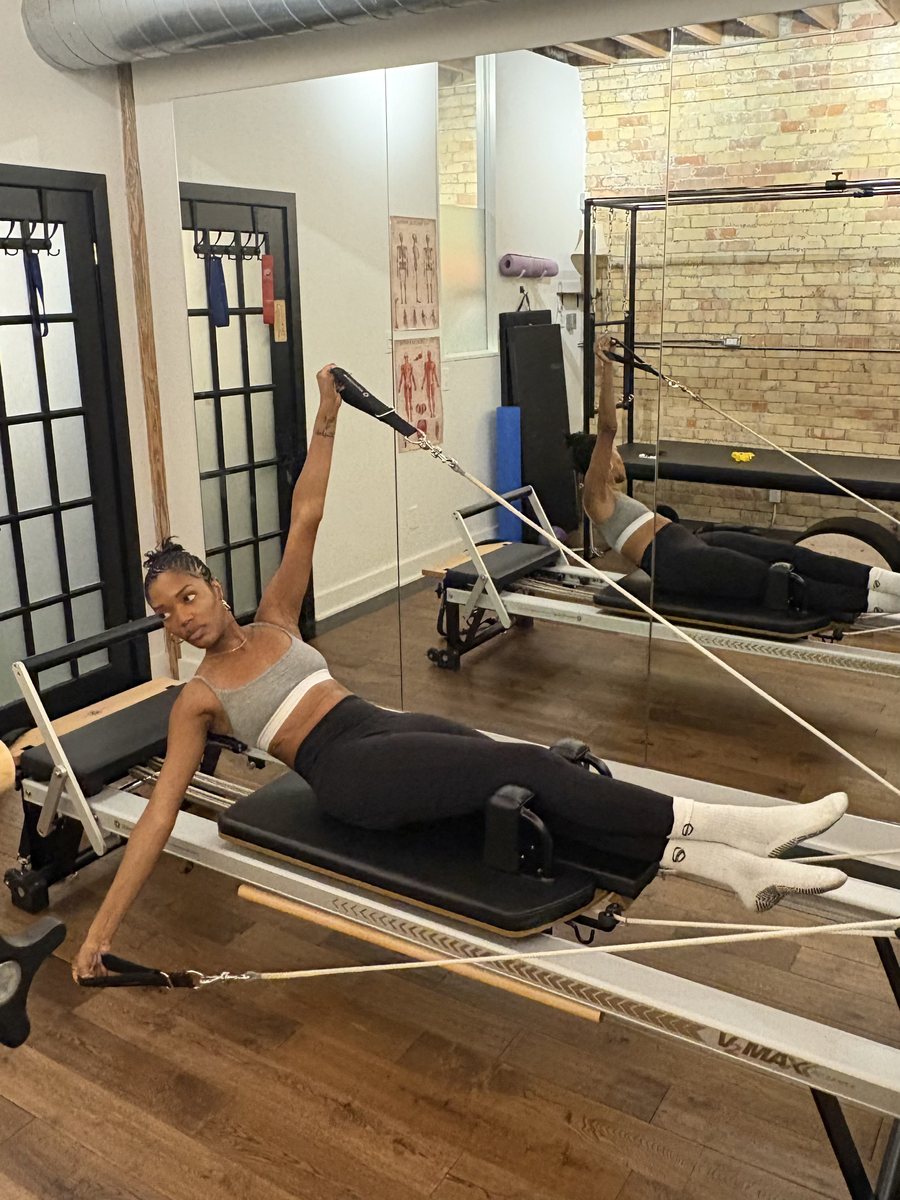 Ruqayyah on the reformer in a long side stretch, one arm reaching overhead